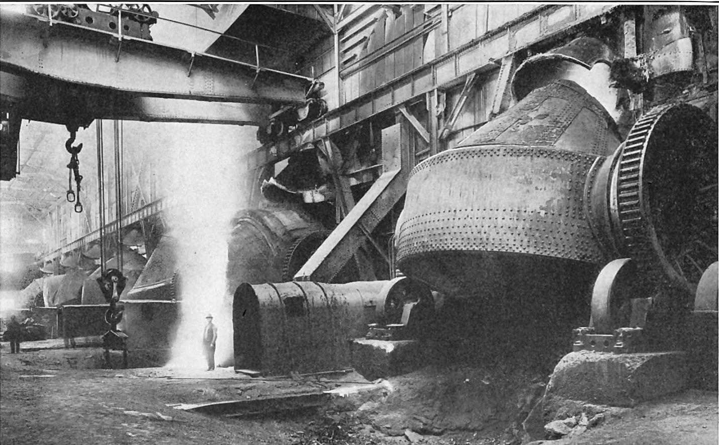 Huge barrel-shaped copper converters under a crane, a worker standing small beside a shower of sparks