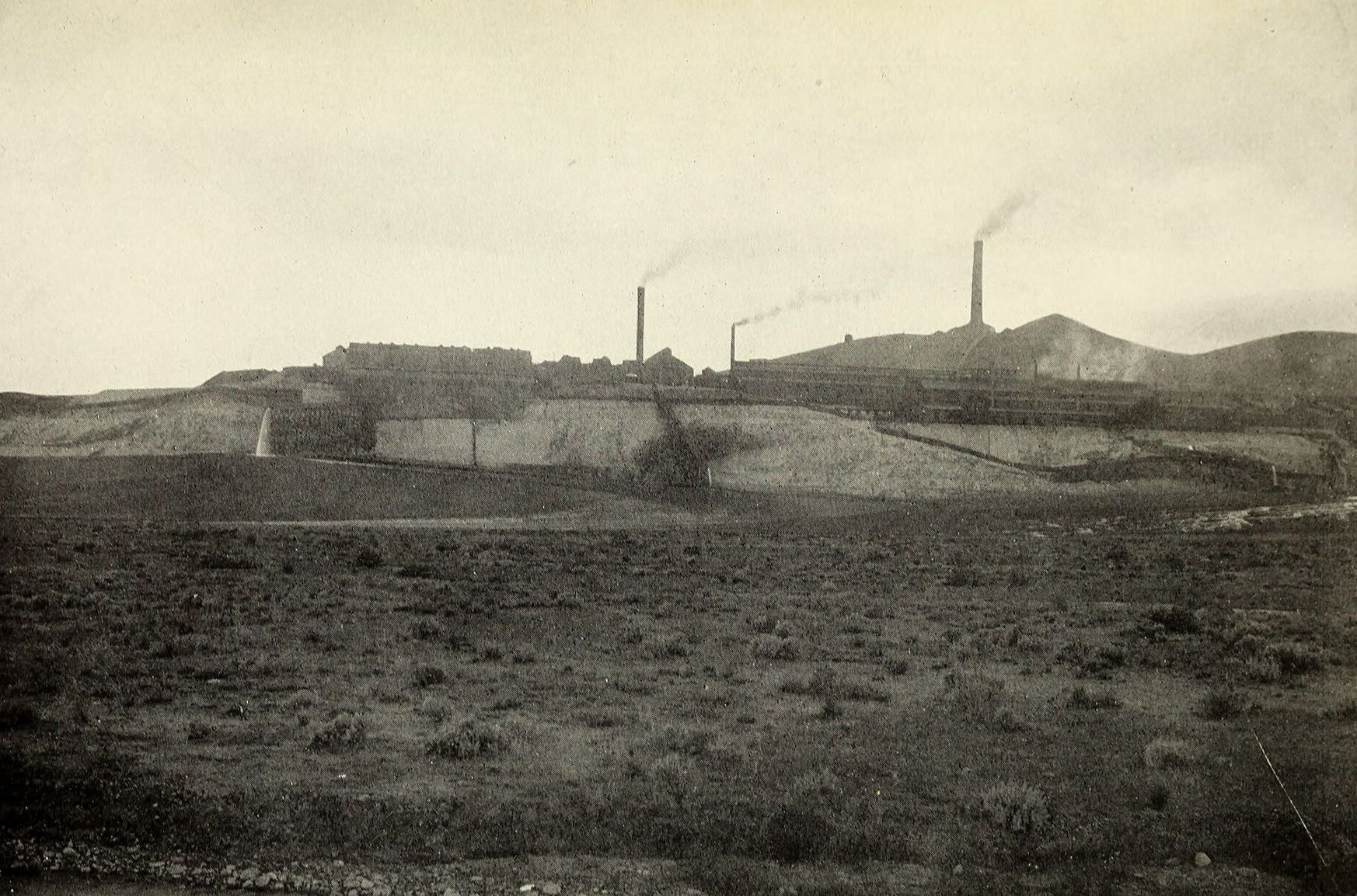Distant smelter stacks smoking on a hill above a flat, dry valley floor, 1908
